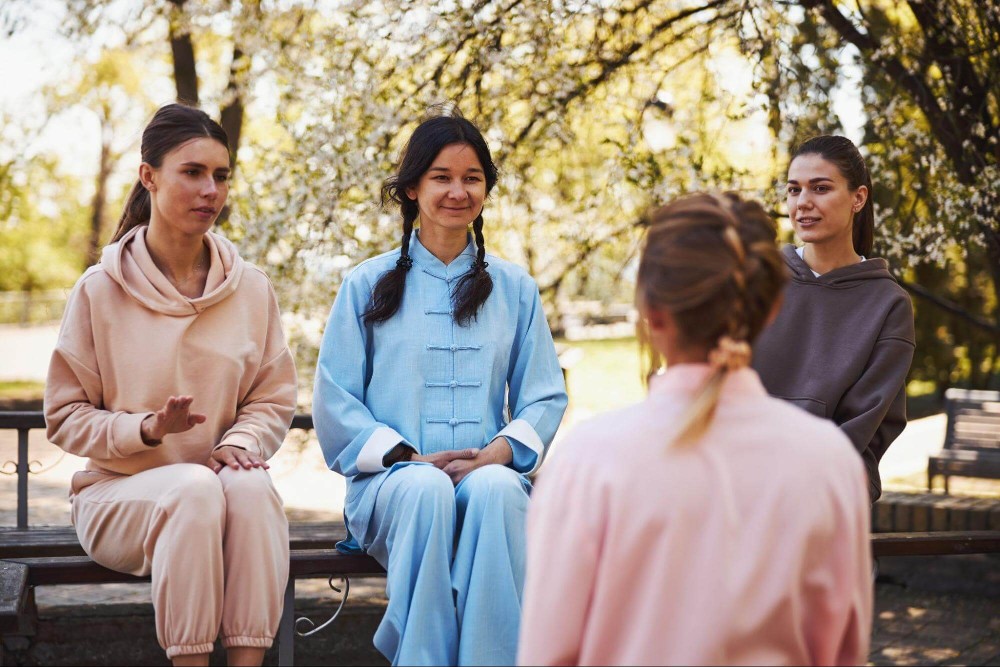 Four women in robes sitting together on a wooden bench, smiling and enjoying each other's company.