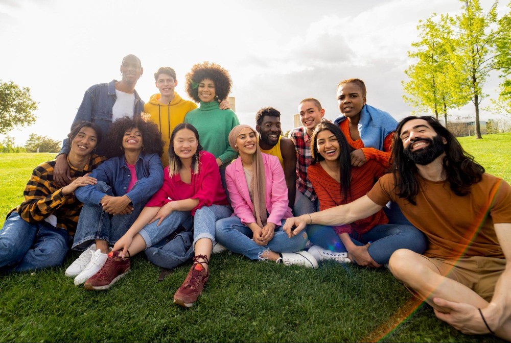 A diverse group of young people sitting on the grass, enjoying a sunny day together in a park setting.