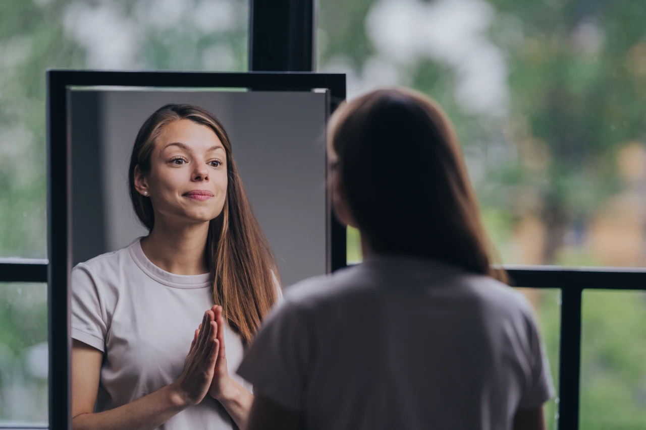 A woman stands indoors, looking at herself in a mirror with her hands pressed together, wearing a white t-shirt—a quiet moment of self acceptance with ADHD.