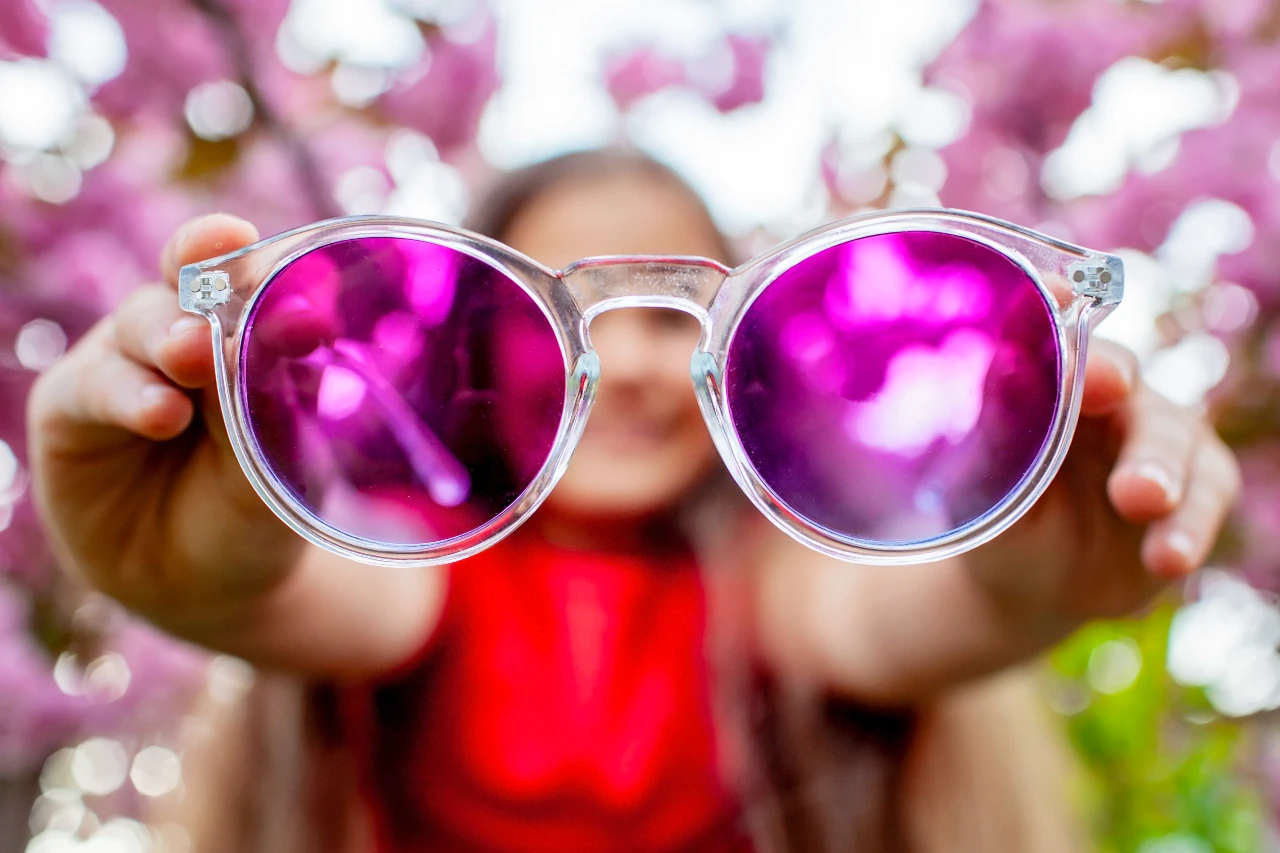 A girl joyfully holds a pair of pink sunglasses, highlighting their vibrant color and trendy style.