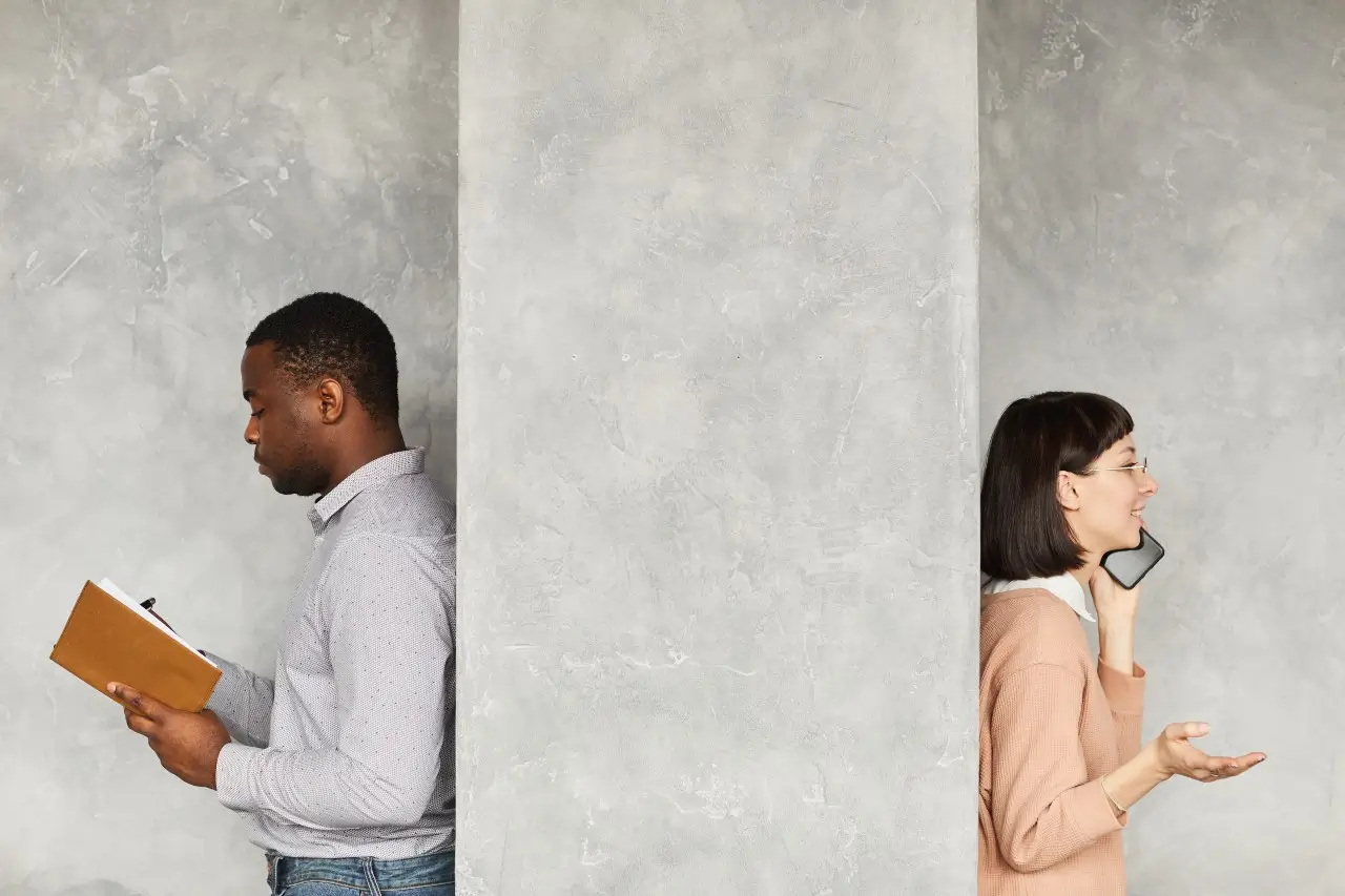 businessman reading his note pad with businesswoman talking on the mobile phone they standing back to back behind the white wall