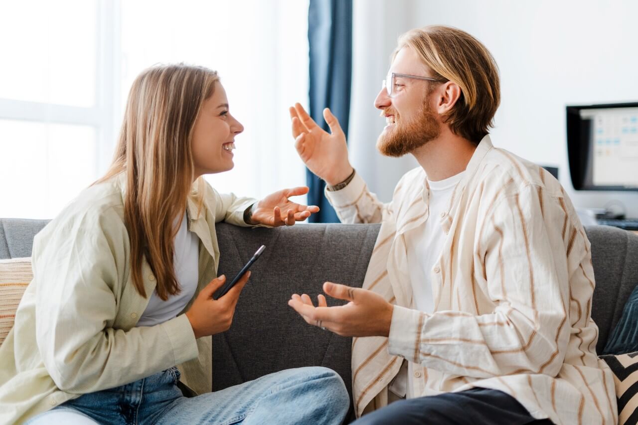 A man and woman sit on a couch, engaged in conversation, smiling and looking at each other.