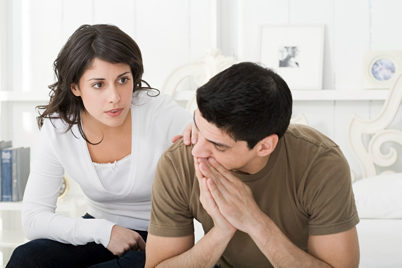 A man and woman sitting together on a bed, engaged in conversation and sharing a moment of intimacy.