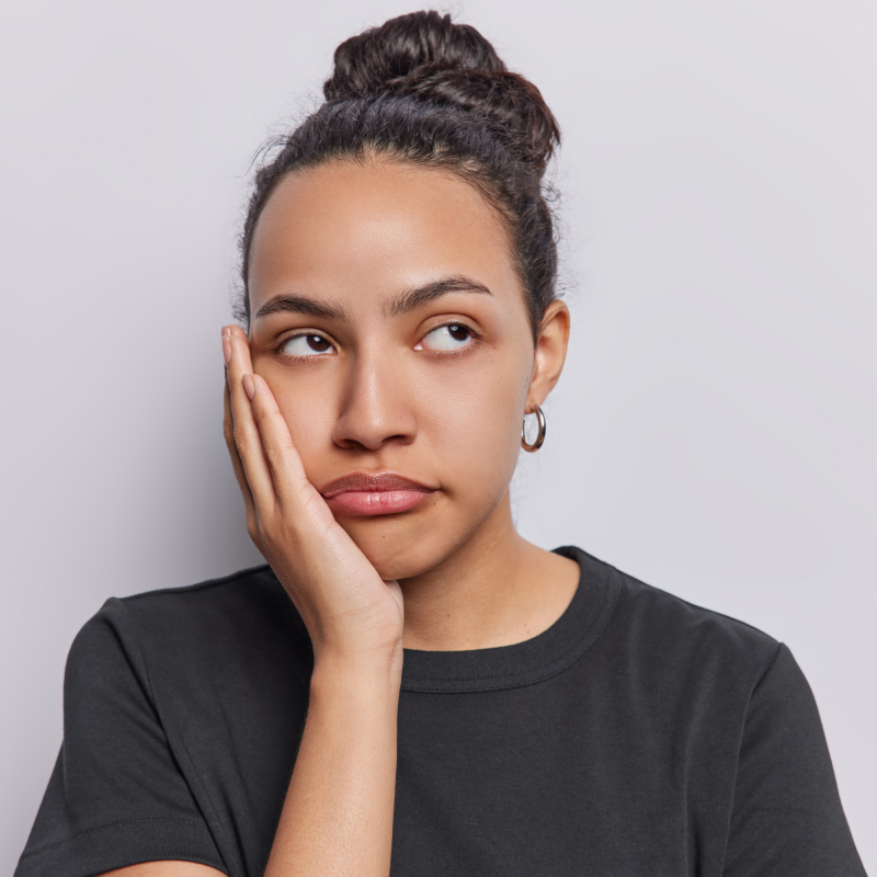 Horizontal shot of upset woman keeps hand on cheek feels bored concentrated aside dressed in
