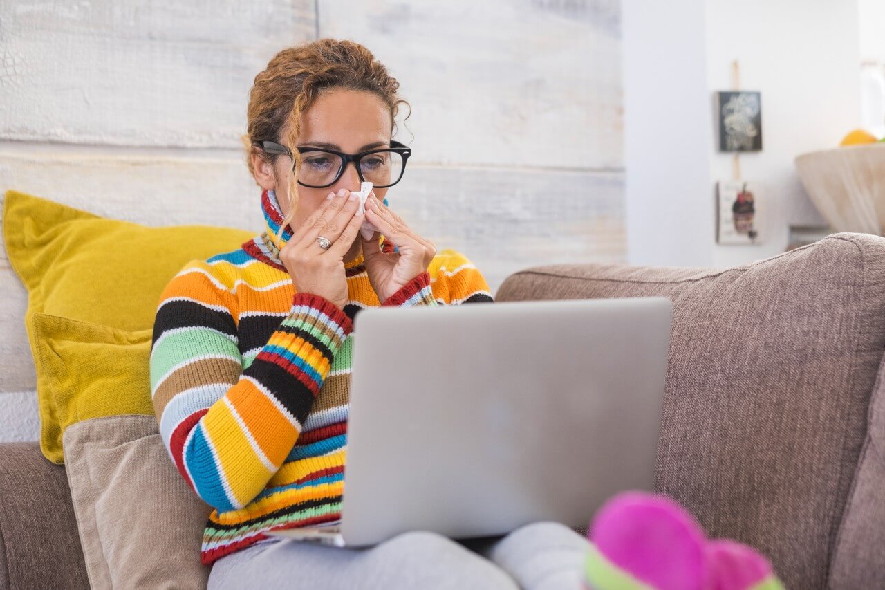 A person in a colorful striped sweater sitting on a couch, blowing their nose while searching symptoms on a laptop, illustrating the link between anxiety and googling health concerns.