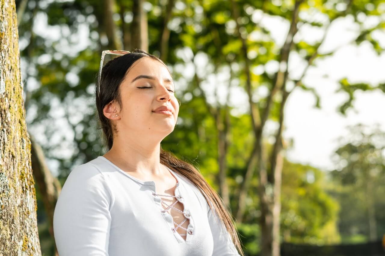 A woman standing outdoors with eyes closed, breathing calmly against a tree, symbolizing recovery and mindfulness after overcoming health anxiety.