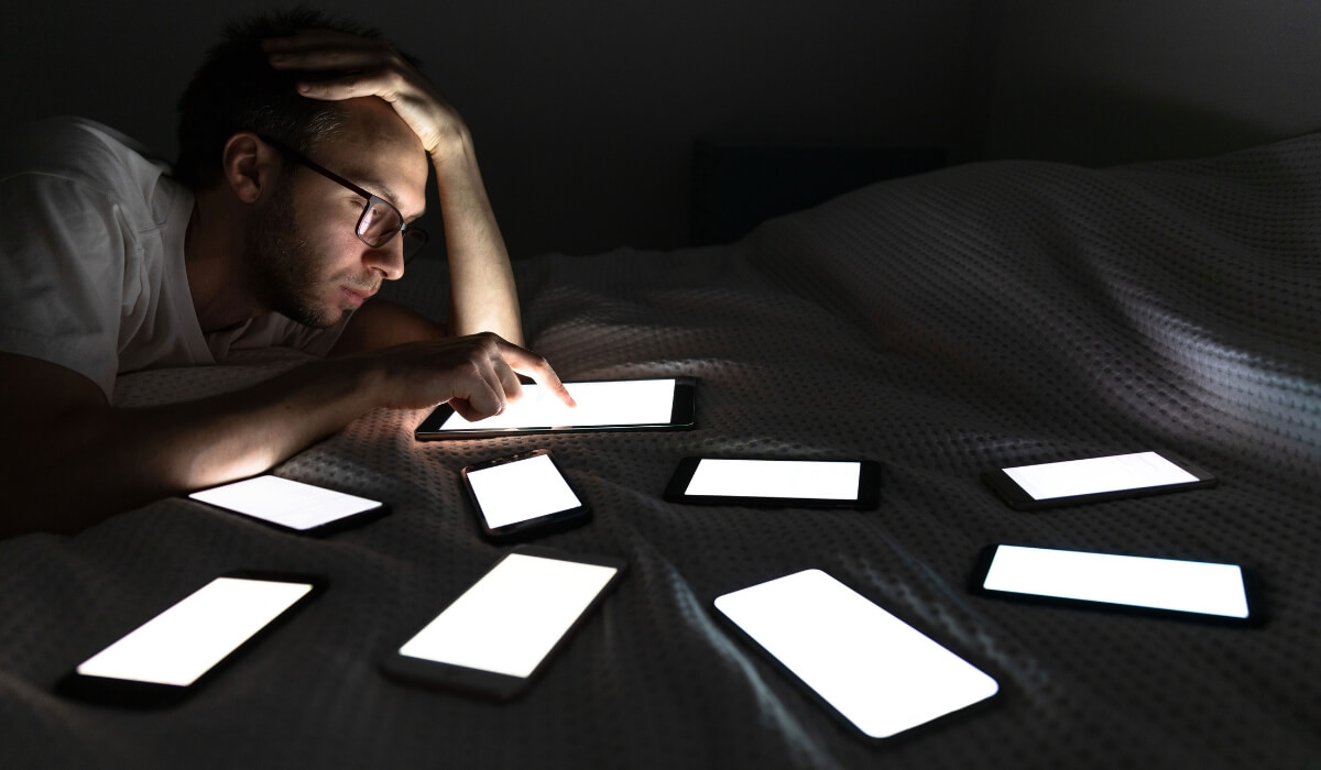 A man uses a tablet in bed at night, surrounded by bright screens symbolizing digital overload. Image used by Get Reconnected Psychotherapy and Counselling Services. Contact info@getreconnected.ca.