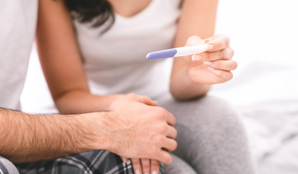 A woman shows a pregnancy test to her partner in bed, with a positive result indicated. The couple is sitting up in bed, and the woman is holding the test up in front of her partner, cropped