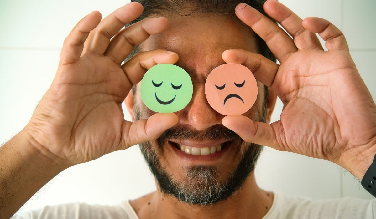 A man playfully covers his eyes with happy and sad emoji icons. Image used by Get Reconnected Psychotherapy and Counselling Services. Contact info@getreconnected.ca.