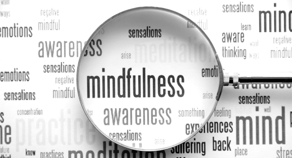 The image presents a magnifying glass emphasizing the term "mindfulness" within a word cloud of related concepts like awareness and meditation, rendered in a monochrome palette.