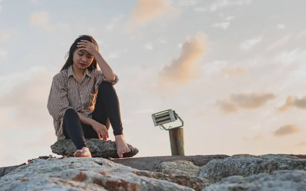 A woman sitting on a rock, appearing distressed as she rests her head in her hands.