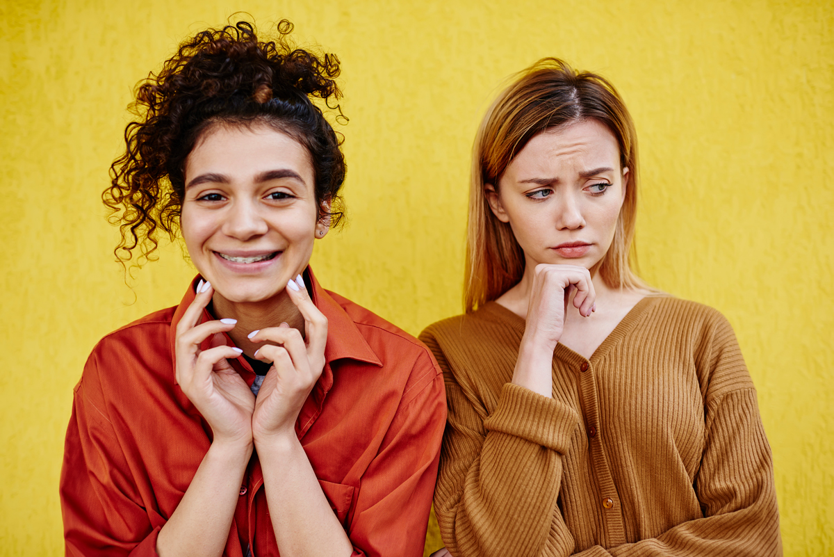 dangers-of-people-pleasing Two women standing side by side, each with their hands resting on their chin, appearing thoughtful.